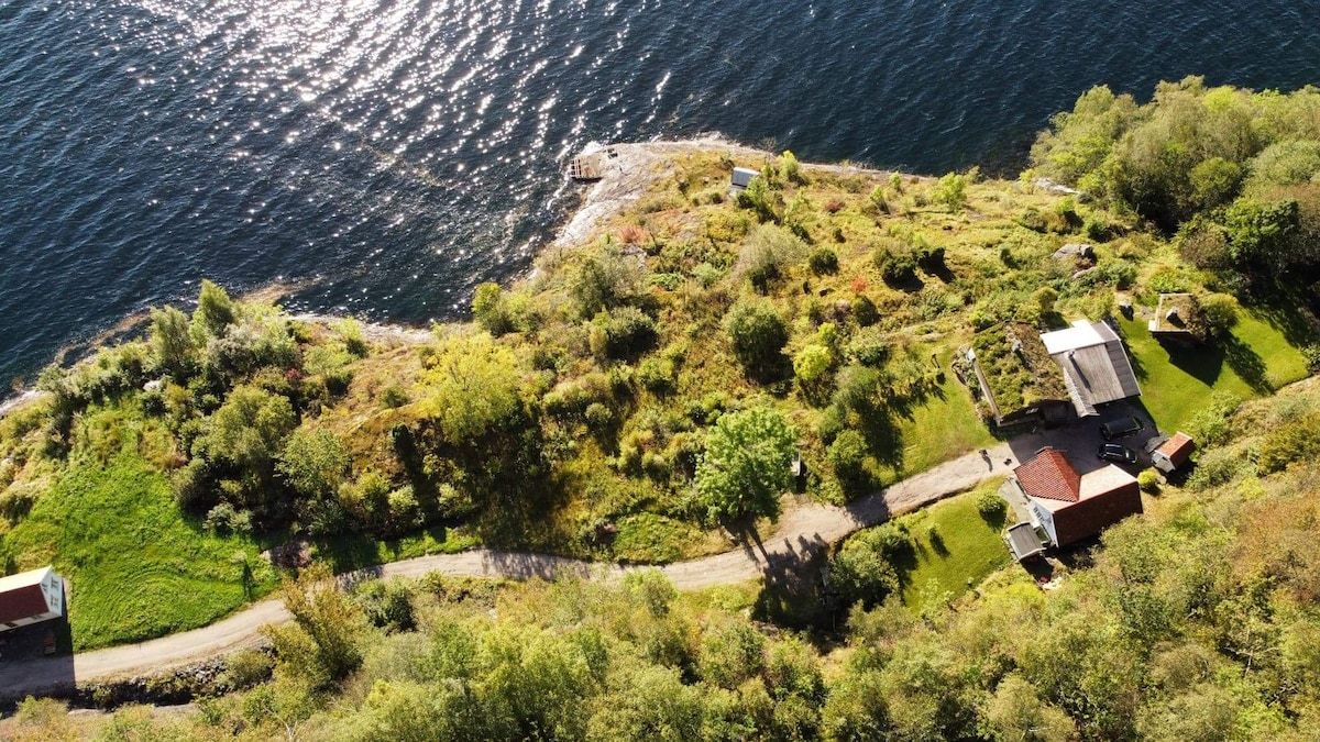 An aerial view captures a private landscape featuring lush vegetation and winding pathways alongside a shimmering fjord. The arrangement includes a guest house, garden areas, and outdoor spaces surrounded by nature, with a dock jutting out into the water.
