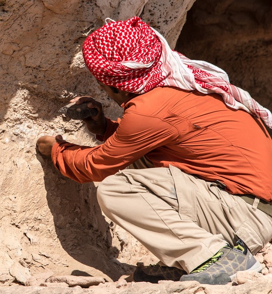 A man is seen crouching against a rocky surface, engaged in the process of chiseling or carving. He is wearing an orange long-sleeve shirt and beige pants, with a traditional red and white patterned scarf wrapped around his head. The natural surroundings feature earthy tones.