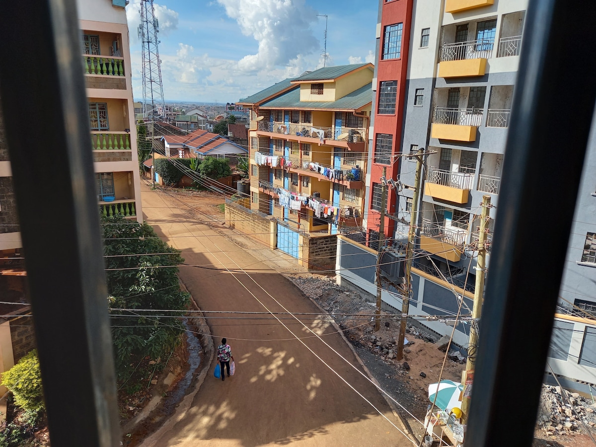 The view from a window showcases a vibrant residential street scene. Colorful apartment buildings are visible, with laundry hanging on balconies. A dirt road runs through the foreground, where a person carries a bag. Power lines and a clear sky add to the urban landscape.