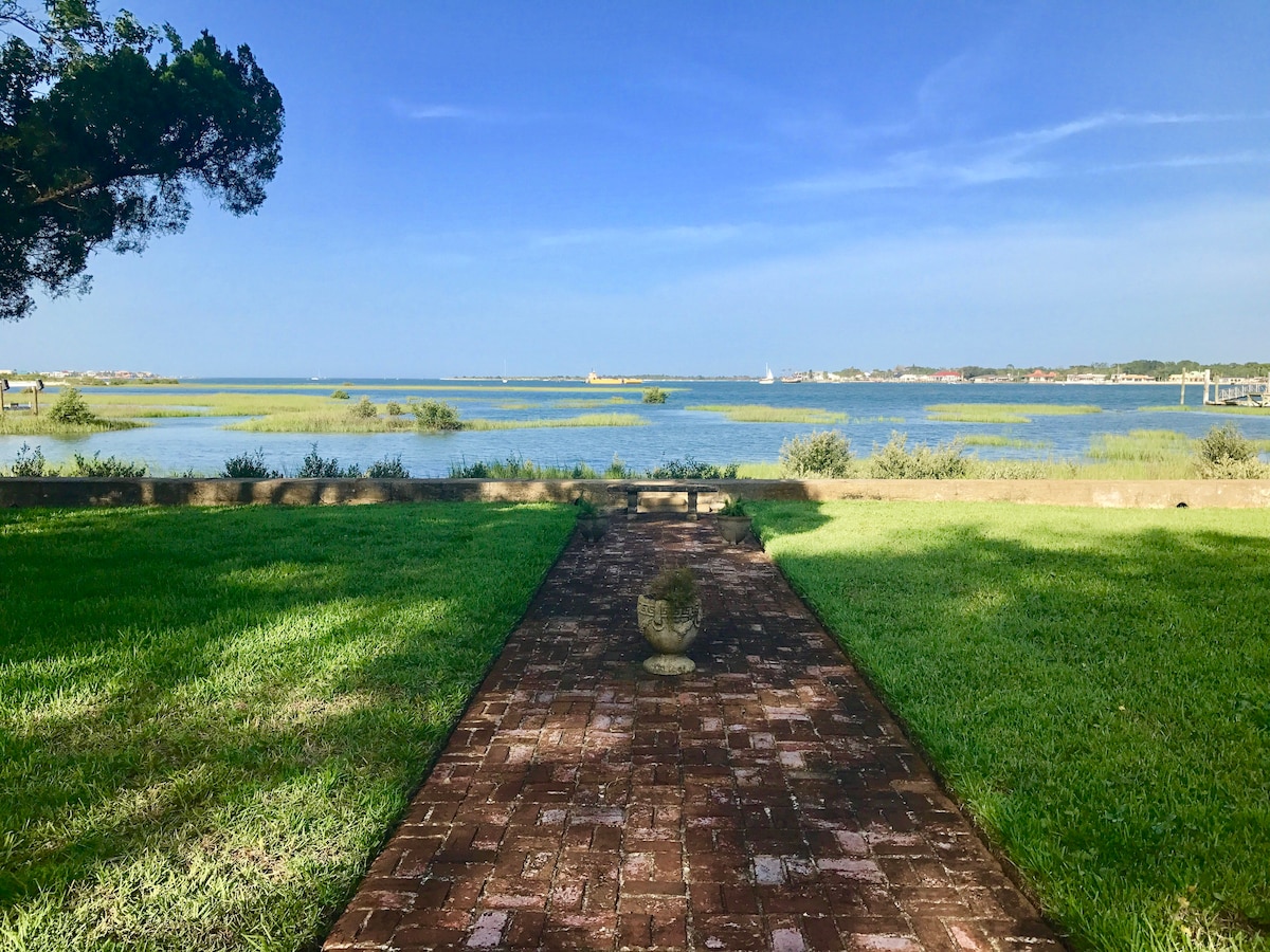 A well-maintained yard features lush green grass leading to a brick path. At the path's end, a stone sculpture is visible, with serene waterfront views of the waterway and distant land. The sky is clear, indicating a bright day.