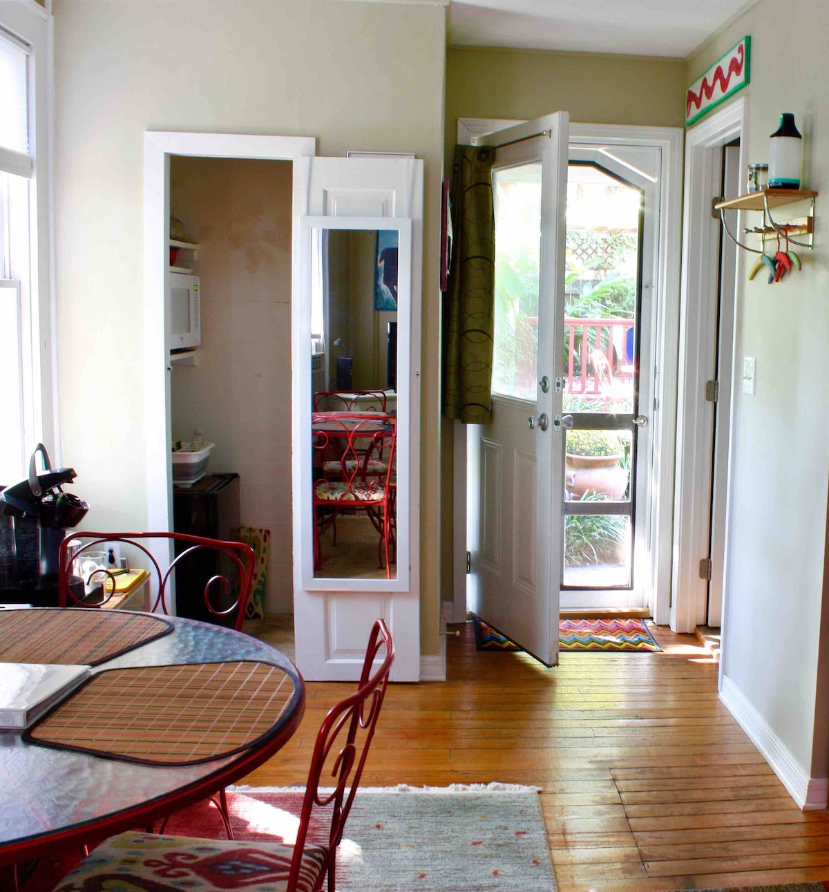 A bright entryway is featured with an open door leading to a small patio. A mirrored cabinet stands adjacent to a cozy dining area with a round table and metal chairs. Wooden floors add warmth to the ambiance, complemented by natural light from nearby windows.