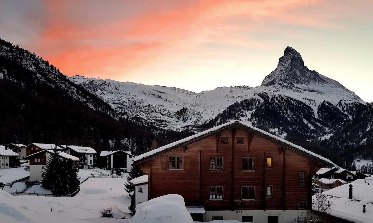 A wooden chalet is framed against a dramatic sunset, with soft pink and orange hues lighting up the sky. The snow-covered landscape extends towards the mountains in the background, prominently featuring a distinctive peak. Nearby buildings are partially visible under the snow.