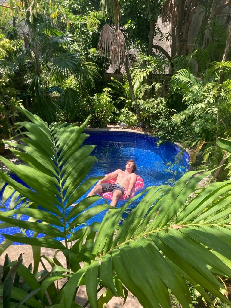 A tranquil pool is surrounded by lush green vegetation. Bright blue water reflects the sunlight, while a circular pink float rests on the surface. Tropical plants frame the scene, creating a secluded, serene atmosphere in the midst of nature.