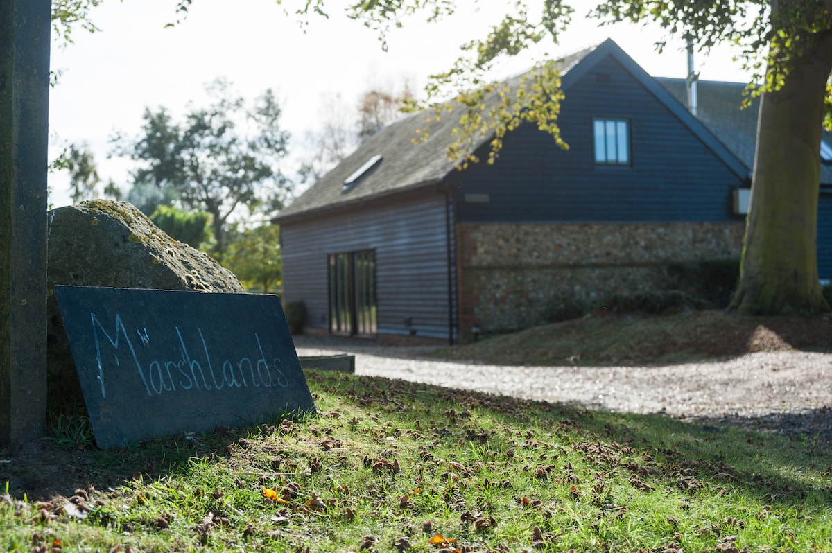 A welcoming sign for Marshlands is positioned on the lush green grass, accompanied by a natural stone. The purpose-built chalet is visible in the background, featuring a dark exterior and expansive glass doors, nestled within a scenic environment.