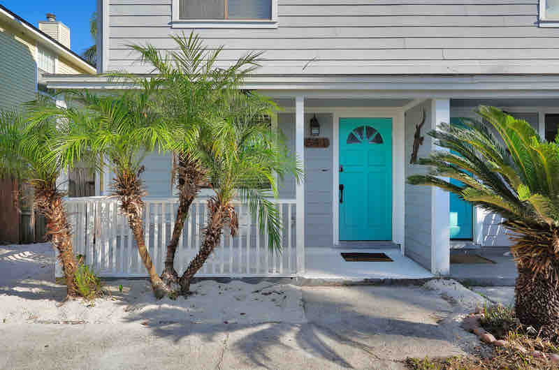 A charming entrance features a bright turquoise front door, surrounded by lush palm trees. The welcoming porch provides a shaded area, with sandy ground indicating a beachy environment. The home's light grey siding complements the outdoor greenery.