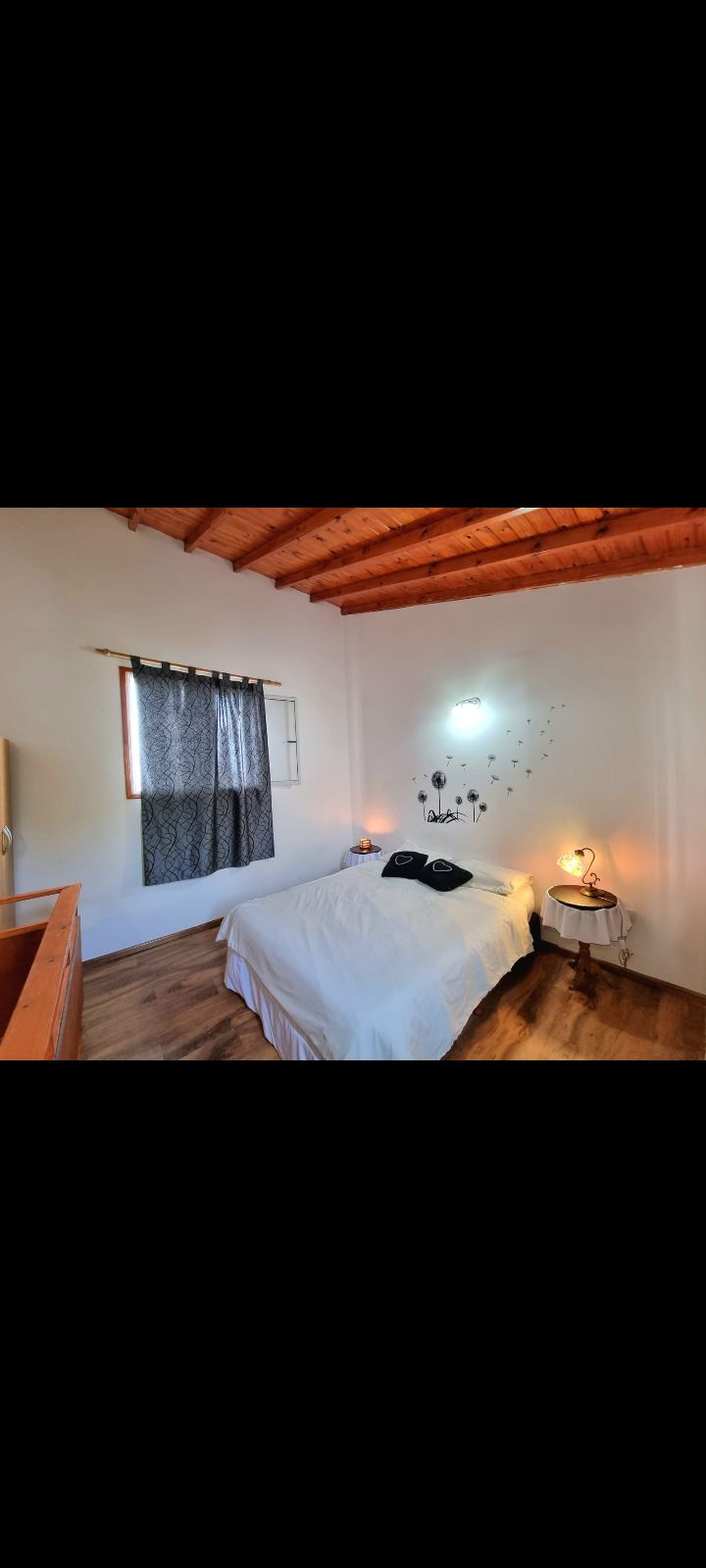 A bright sleeping area features a spacious bed covered with a light comforter. A small bedside table holds a lamp, and patterned curtains frame the window, allowing natural light to filter in. A decorative wall adds character to the warm wooden ceiling.