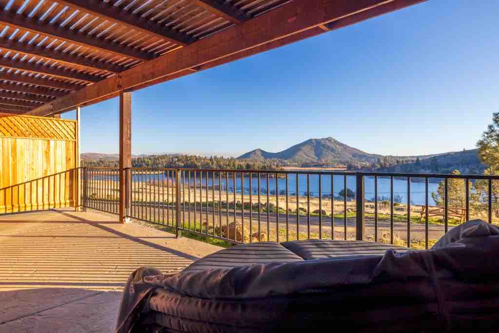 A covered terrace offers a view of the tranquil lake, surrounded by trees and rolling hills. The space features a dark lounge chair and a railing that outlines the area, while shadows from the pergola create a striped pattern on the floor.