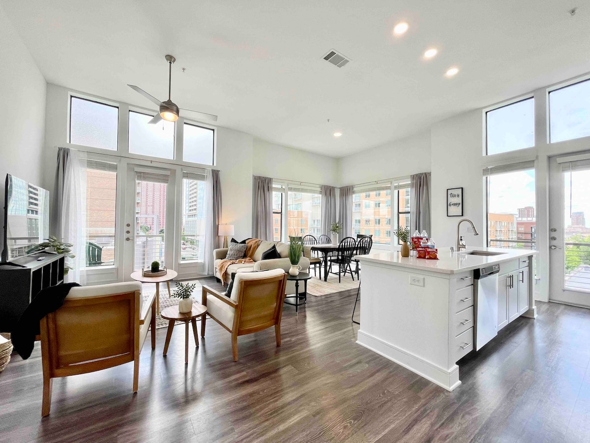 A spacious living area features large windows that allow natural light to illuminate the room. A comfortable seating arrangement of stylish chairs and a sofa overlooks a dining area with a round table. A modern kitchen with white cabinetry is partially visible, enhancing the open layout.