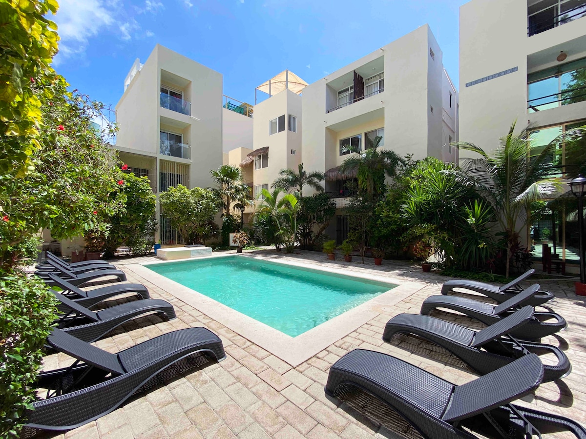 A serene swimming pool is surrounded by lush greenery, featuring several modern lounge chairs positioned on textured stone paving. The bright blue sky reflects into the clear water, creating a calming and inviting area for relaxation.