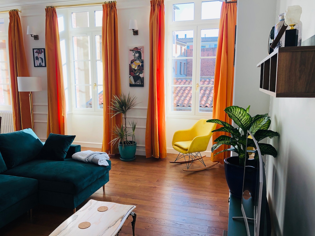 A bright living area features large windows adorned with orange curtains, allowing ample natural light. A plush teal sofa is complemented by a vibrant yellow rocking chair. Decorative plants add a touch of greenery, enhancing the welcoming atmosphere of the space.