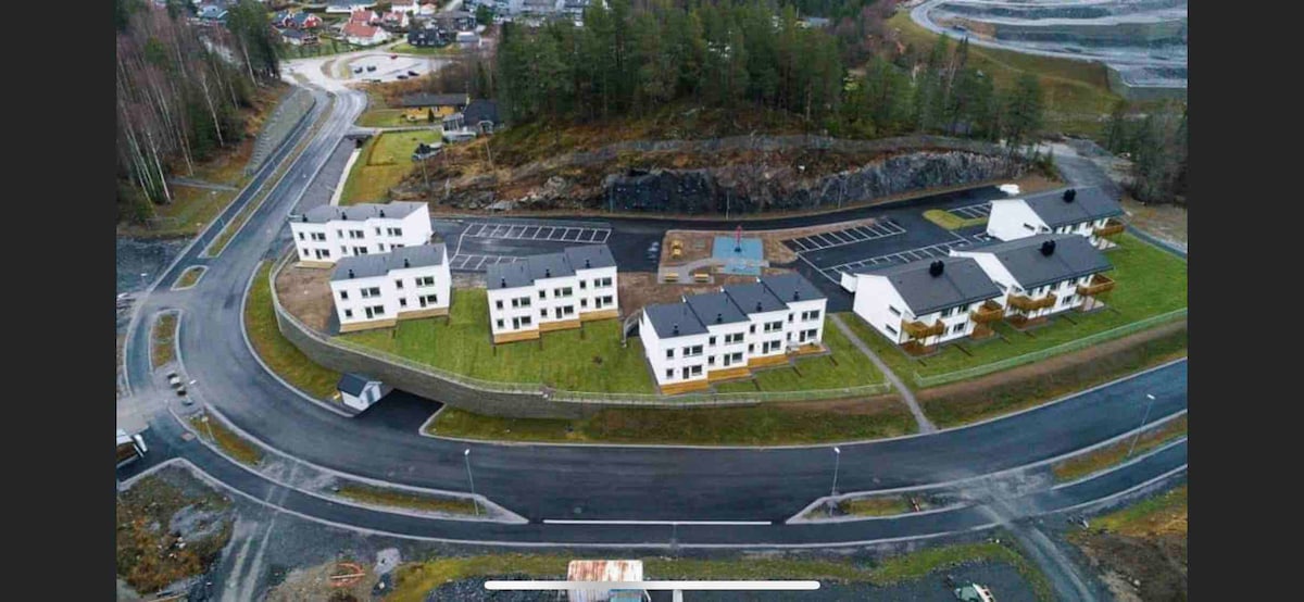 An aerial view showcases a cluster of modern white buildings arranged in a circular layout, surrounded by grassy areas. Nearby, a parking lot is visible, along with pathways leading to outdoor spaces. The backdrop features a rocky landscape and tree line.