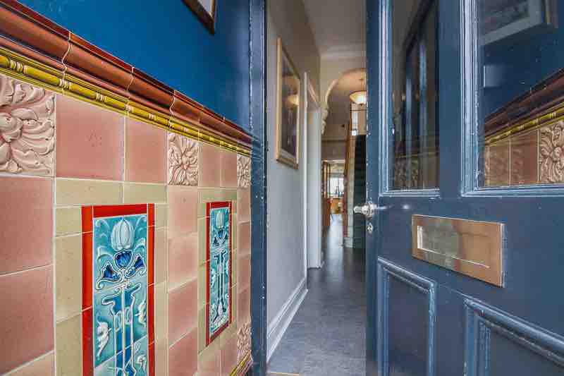 An inviting entrance displays intricate tilework alongside the doorframe. The hallway leads to spacious areas, showcasing glimpses of light and the character of the home. Decorative elements are present, enhancing the overall charm of the entryway.