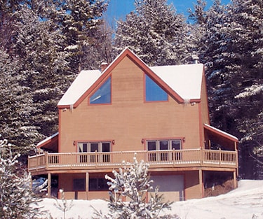 The exterior of a two-story vacation home is displayed, surrounded by snow-covered trees. A wrap-around deck extends around the home, featuring large windows that allow natural light to enter. The roof is sloped, and the overall structure is designed for a spacious and inviting appearance.