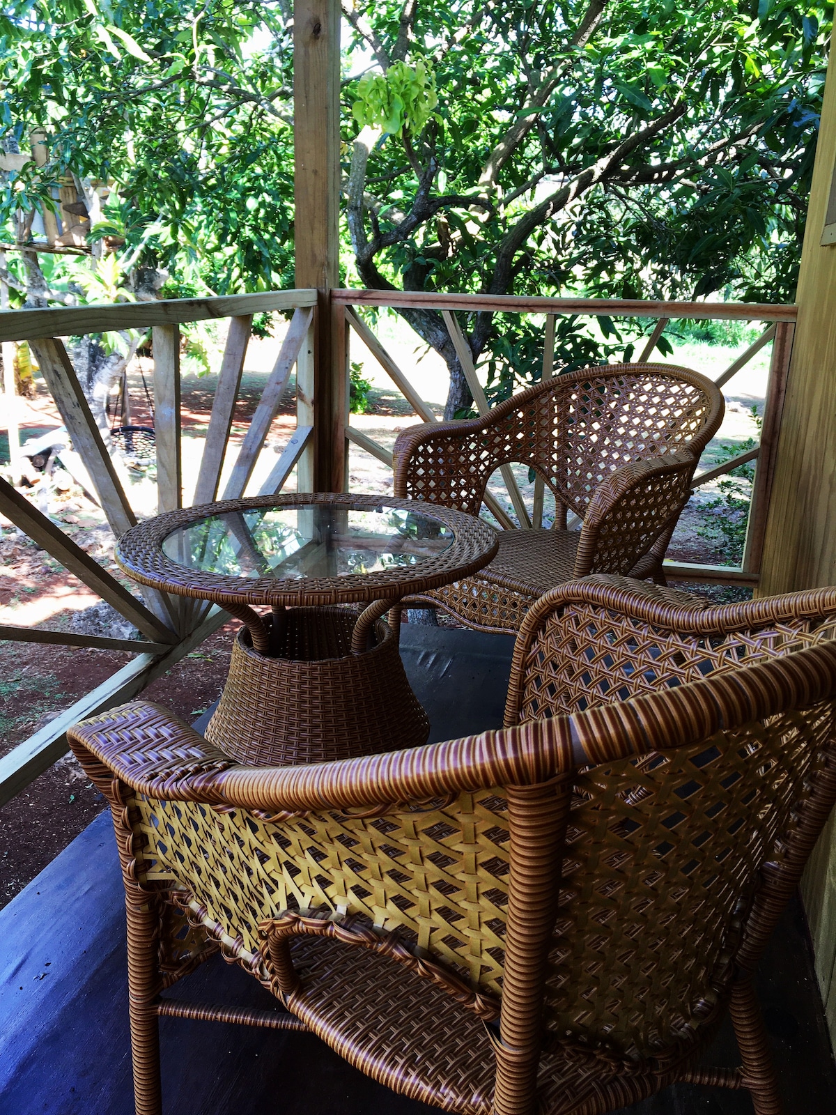 A shaded private veranda features a small glass-topped table surrounded by two woven chairs. The area is framed by wooden railings and overlooks a lush garden with tree branches visible in the background.