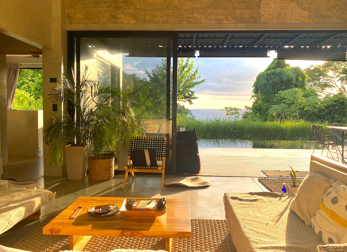 The interior space features an open layout with large glass doors that provide a view of the natural surroundings and ocean. A comfortable seating area is present, complemented by a wooden coffee table. A pool and lush greenery are visible outside, enhancing the tranquil ambiance.