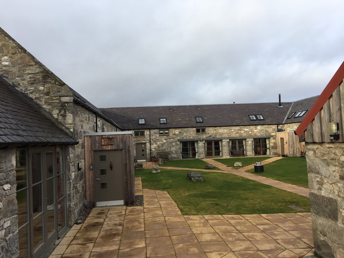 A spacious courtyard is surrounded by stone buildings featuring a combination of slate and wooden rooftops. The well-maintained grass area contains stone pathways, garden furniture, and a seating area, inviting relaxation in a serene outdoor setting.