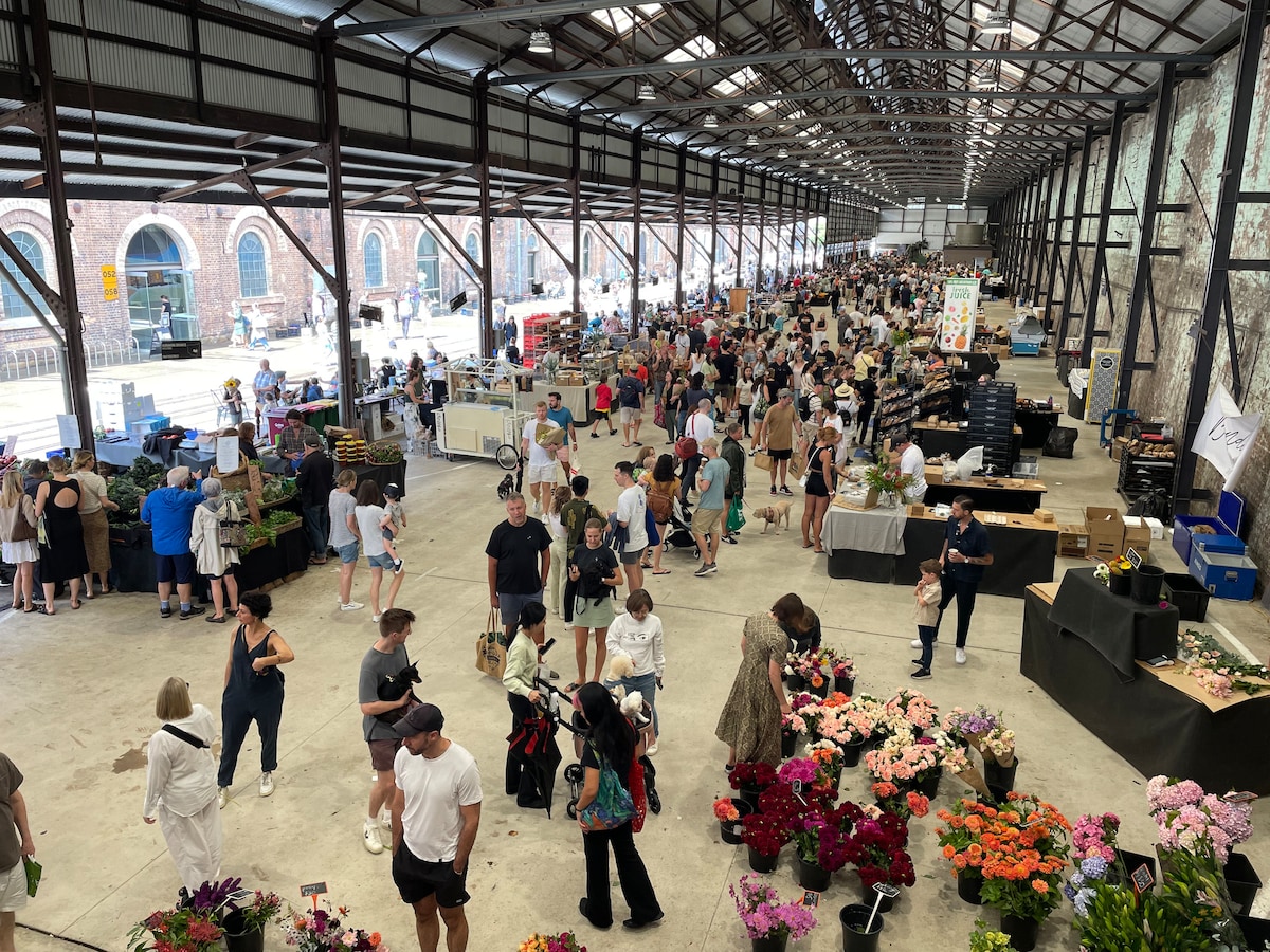 A bustling indoor market scene is captured, showcasing a wide array of market stalls filled with fresh produce and flowers. Vendors and shoppers are seen navigating through the space, which is characterized by high ceilings and a rustic industrial aesthetic.