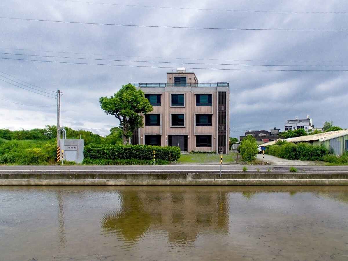 A modern four-story building is situated alongside a quiet road, with large windows reflecting the surrounding greenery. A gently flowing canal runs in the foreground, creating a peaceful atmosphere. Clouds fill the sky, hinting at a calm day.