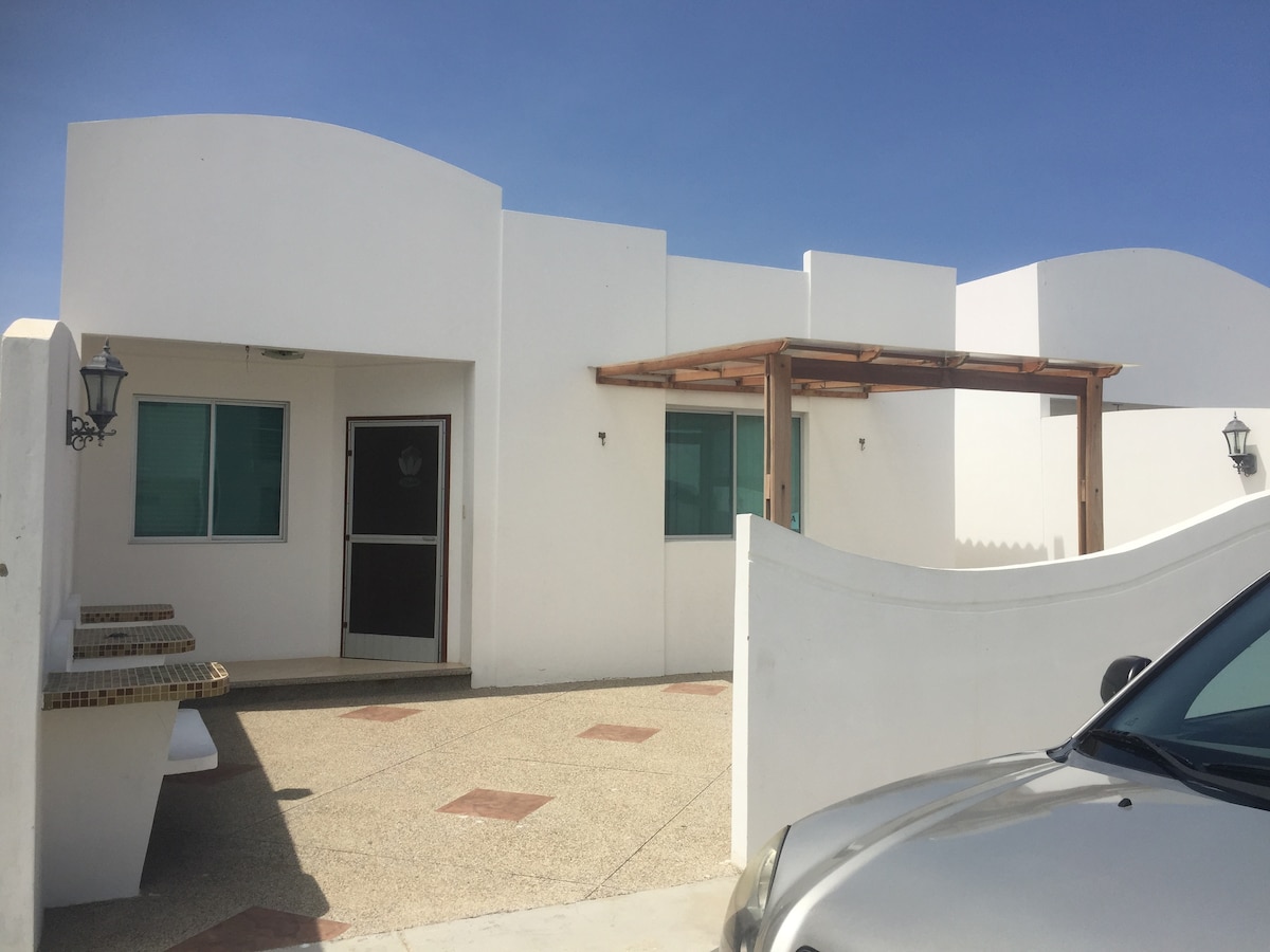 A modern white villa is featured, showcasing a covered entryway supported by wooden beams. The façade includes large windows, a decorative lantern, and a light-colored driveway. A silver car is parked nearby, highlighting the accessible entrance.