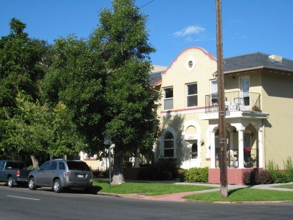 A charming two-story building is depicted, featuring an arched entrance beneath decorative detailing. The building's exterior is complemented by lush green trees and manicured lawns. Balconies are visible on the upper level, and parked cars line the street, highlighting the suburban setting.