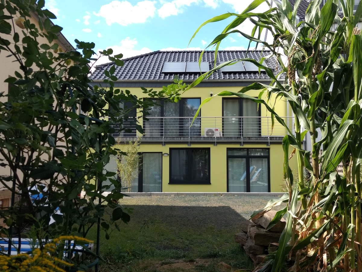 The exterior of the building features a contemporary design with a bright yellow facade. Large glass windows are complemented by a balcony, while solar panels are visible on the roof. Surrounding greenery creates a natural frame, enhancing the inviting atmosphere.