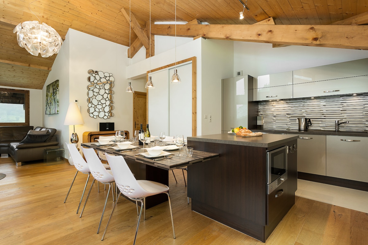 An open-plan kitchen and dining area is presented, featuring a wooden dining table set for six. Modern appliances, including an oven and dishwasher, complement the sleek cabinetry. Natural light illuminates the space through large windows, highlighting the exposed wooden beams and contemporary chandelier.