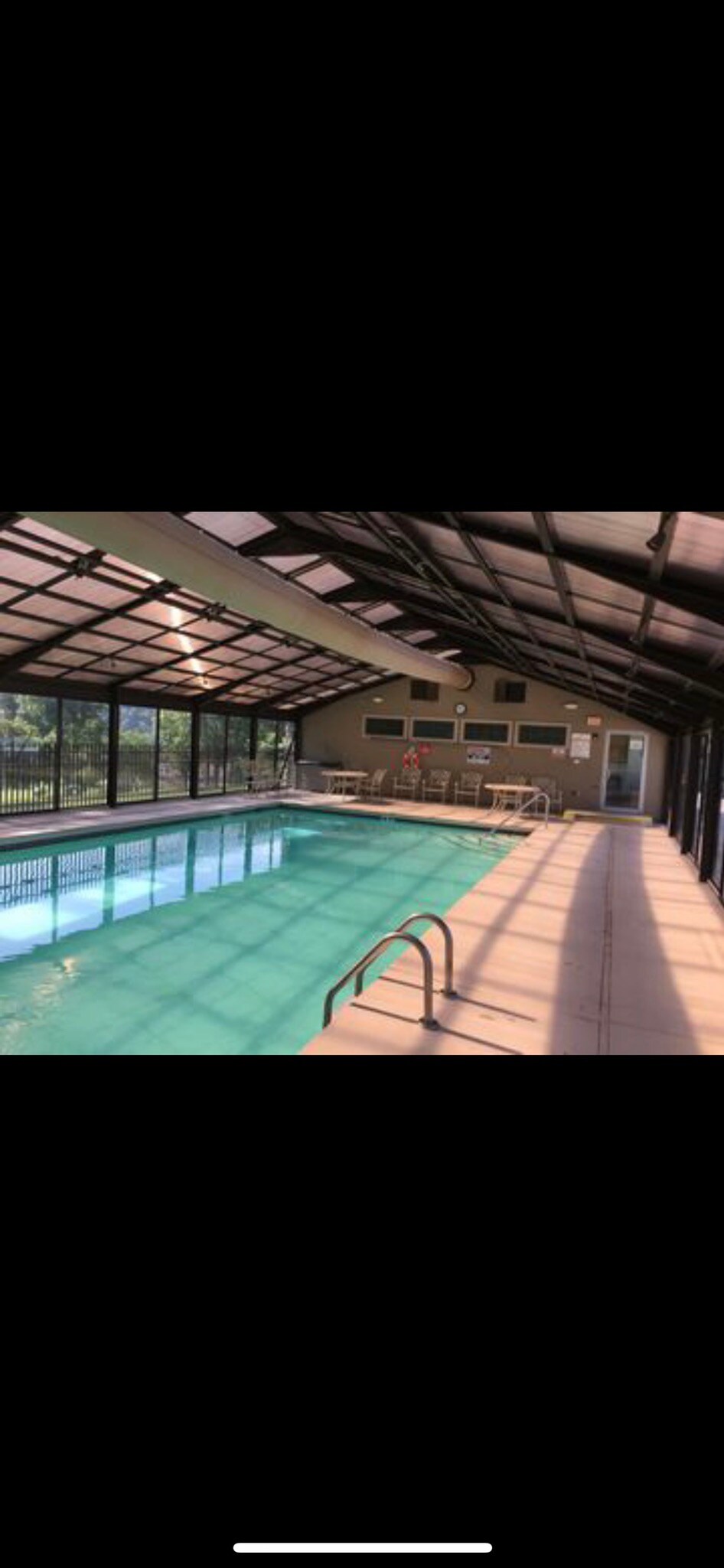 An indoor pool area is showcased, featuring a spacious swimming pool with clear, inviting water. Large glass windows line the walls, allowing natural light to illuminate the area. Poolside chairs are arranged neatly along the edges, enhancing the open and airy feel of the space.