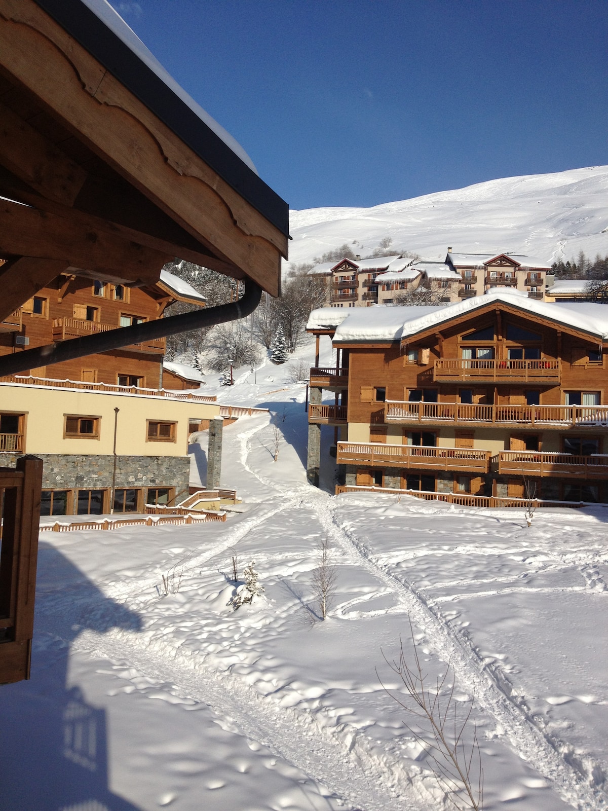 A view of the chalet-style residences nestled in a snowy landscape. The pathway leading towards the buildings, flanked by untouched snow, creates a serene atmosphere. The blue sky overhead complements the snowy scenery, enhancing the winter resort setting.