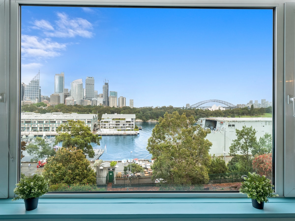 A spacious window frames unobstructed views of Sydney's skyline, featuring the Harbour Bridge and the Opera House. The serene waters below reflect the vibrant cityscape, while green potted plants add a touch of nature to the indoor setting.