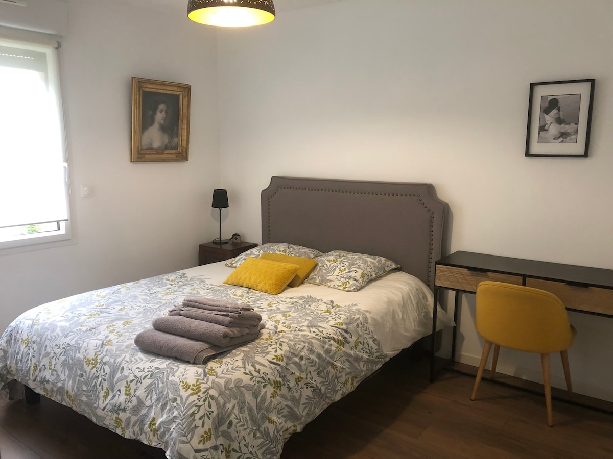 A neatly arranged bedroom features a spacious bed with a floral-patterned quilt and multiple neatly stacked towels. A small bedside table with a lamp sits beside the bed, while a wooden desk with a yellow chair is positioned against the wall. Natural light filters through a nearby window.