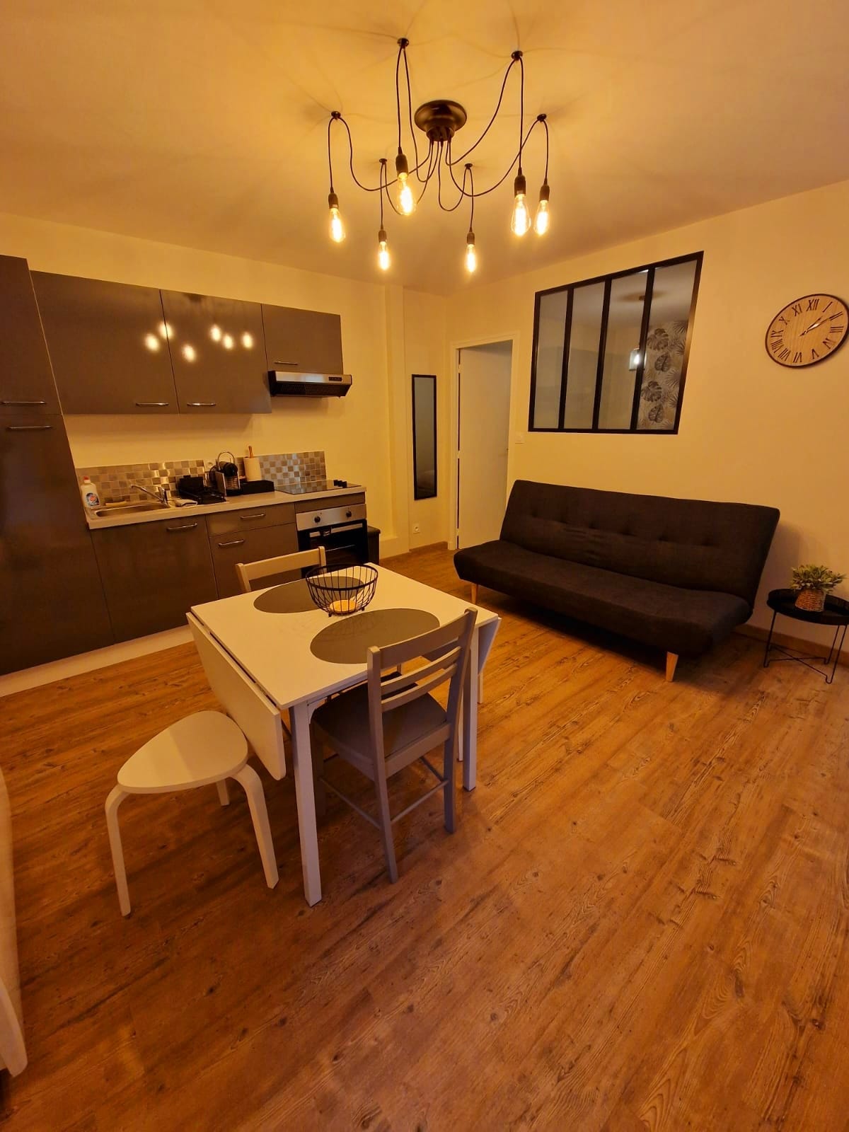The living area features a modern kitchen with sleek cabinets and essential appliances. A dining table is set for four, accompanied by a comfortable sofa and stylish lighting overhead. The warm wooden floor enhances the contemporary design, creating an inviting space for relaxation.