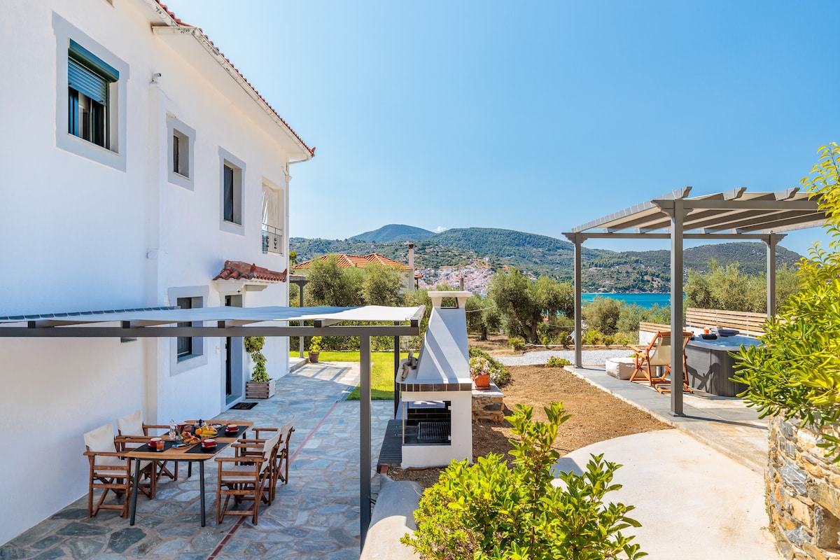An outdoor space features a shaded area with seating arrangements and a dining table, set against a backdrop of distant hills and the sea. A stone path leads to a barbecue area, surrounded by well-maintained greenery and sunlight illuminating the open patio.