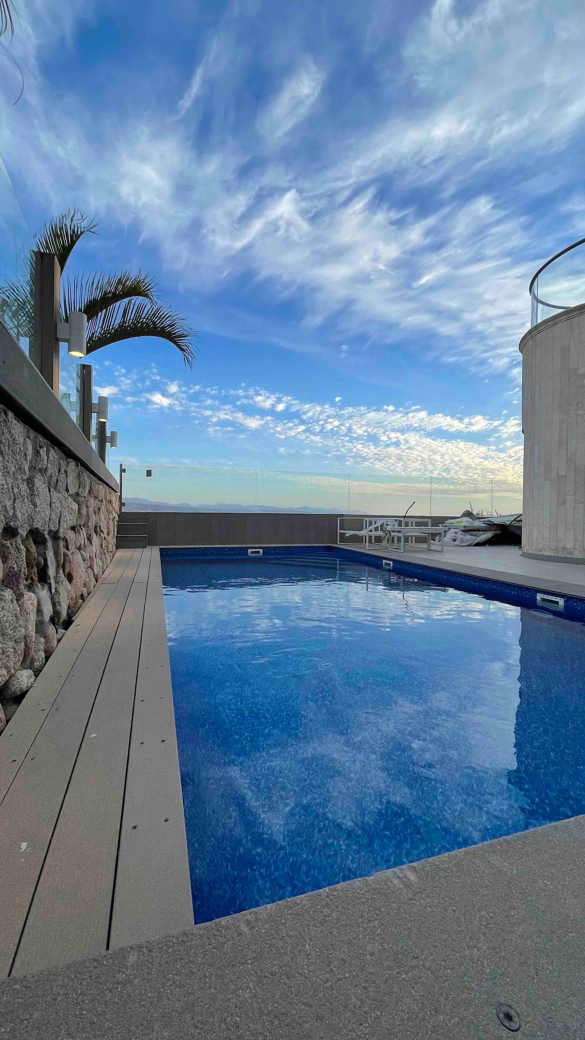 A private swimming pool is surrounded by a smooth stone deck, reflecting clear blue skies filled with wispy clouds. The area features a low stone wall and tropical plants can be seen nearby, enhancing the pool's tranquil setting.