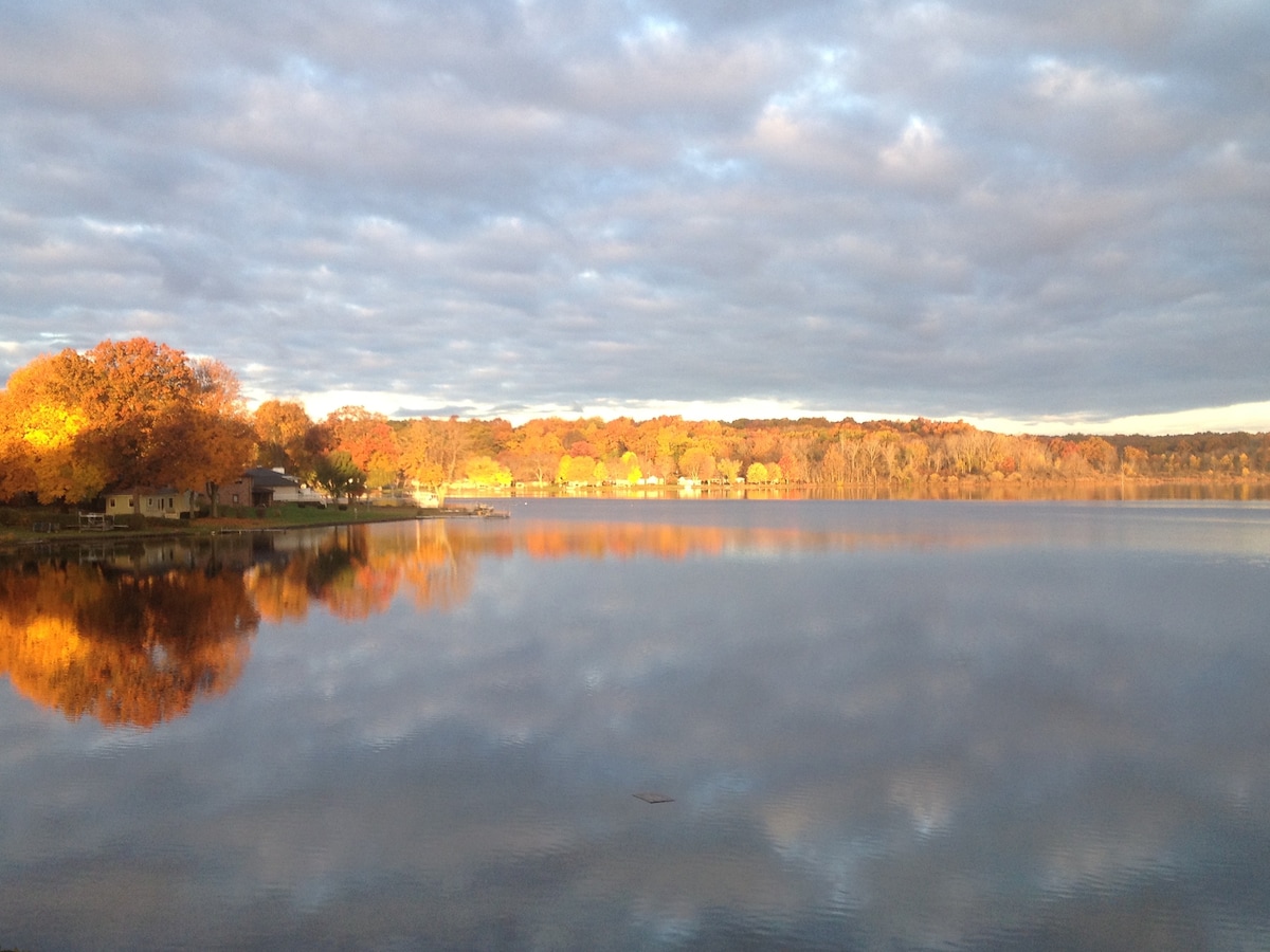 A serene lake scene is captured at sunset, reflecting vibrant autumn foliage along the shoreline. The water's surface mirrors the colorful trees and cloudy sky, creating a peaceful environment. Lakeside houses are visible in the distance, adding to the tranquil atmosphere.