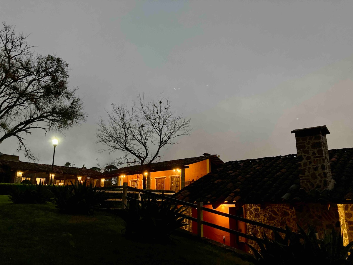 The exterior of the cabañas is softly illuminated at dusk, showcasing rustic stone walls and traditional tile roofs. A tree with sparse branches stands beside the walkway, which is lined with low wooden fencing, enhancing the serene atmosphere of the property.