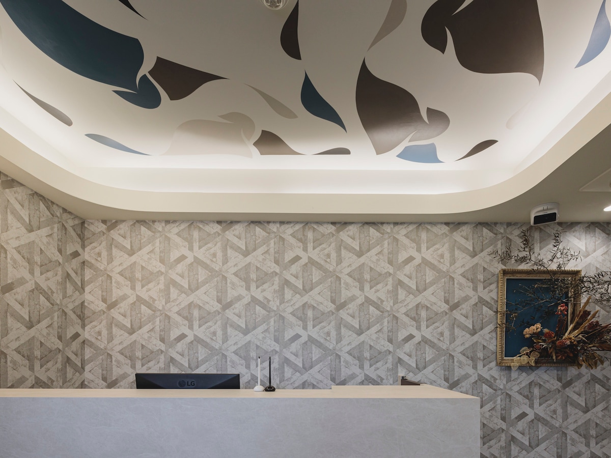 The reception area features a modern design with an artistic ceiling pattern in shades of blue, gray, and beige. A sleek counter is positioned in front, complemented by a decorative wall adorned with a geometric motif and a framed artwork above.