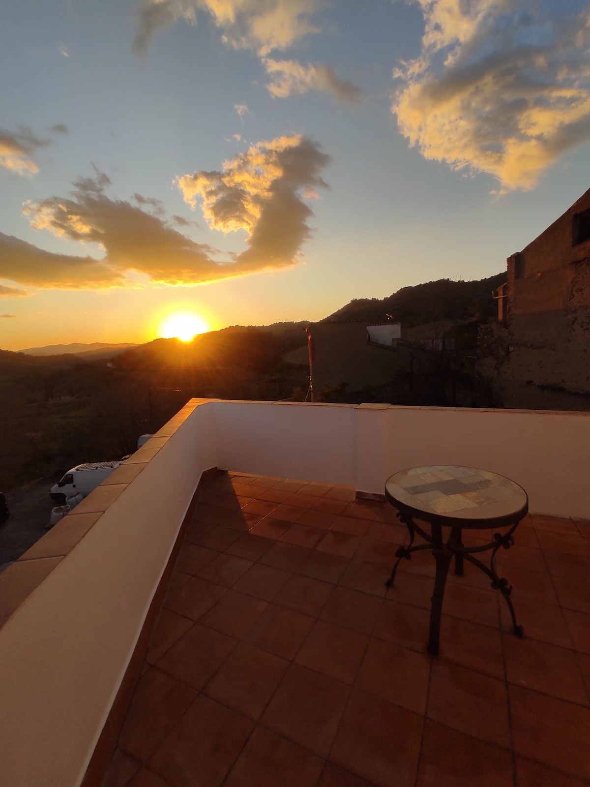 A terrace space offers a scenic view of the sunset, with warm hues illuminating the sky. A small round table is positioned on the terracotta tiled floor, surrounded by natural landscapes and distant hills. The serene atmosphere is enhanced by the mountainous backdrop.
