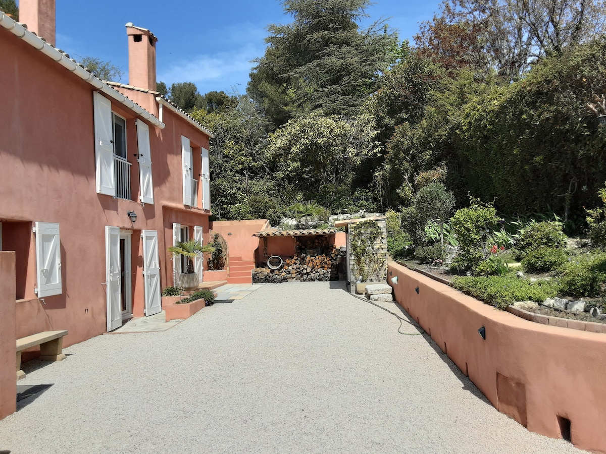 A tranquil exterior view of a pink-stucco house is displayed, surrounded by lush greenery. A gravel pathway leads to the entrance, while a woodpile is neatly stacked against the wall. Bright blue skies enhance the peaceful atmosphere of the garden.
