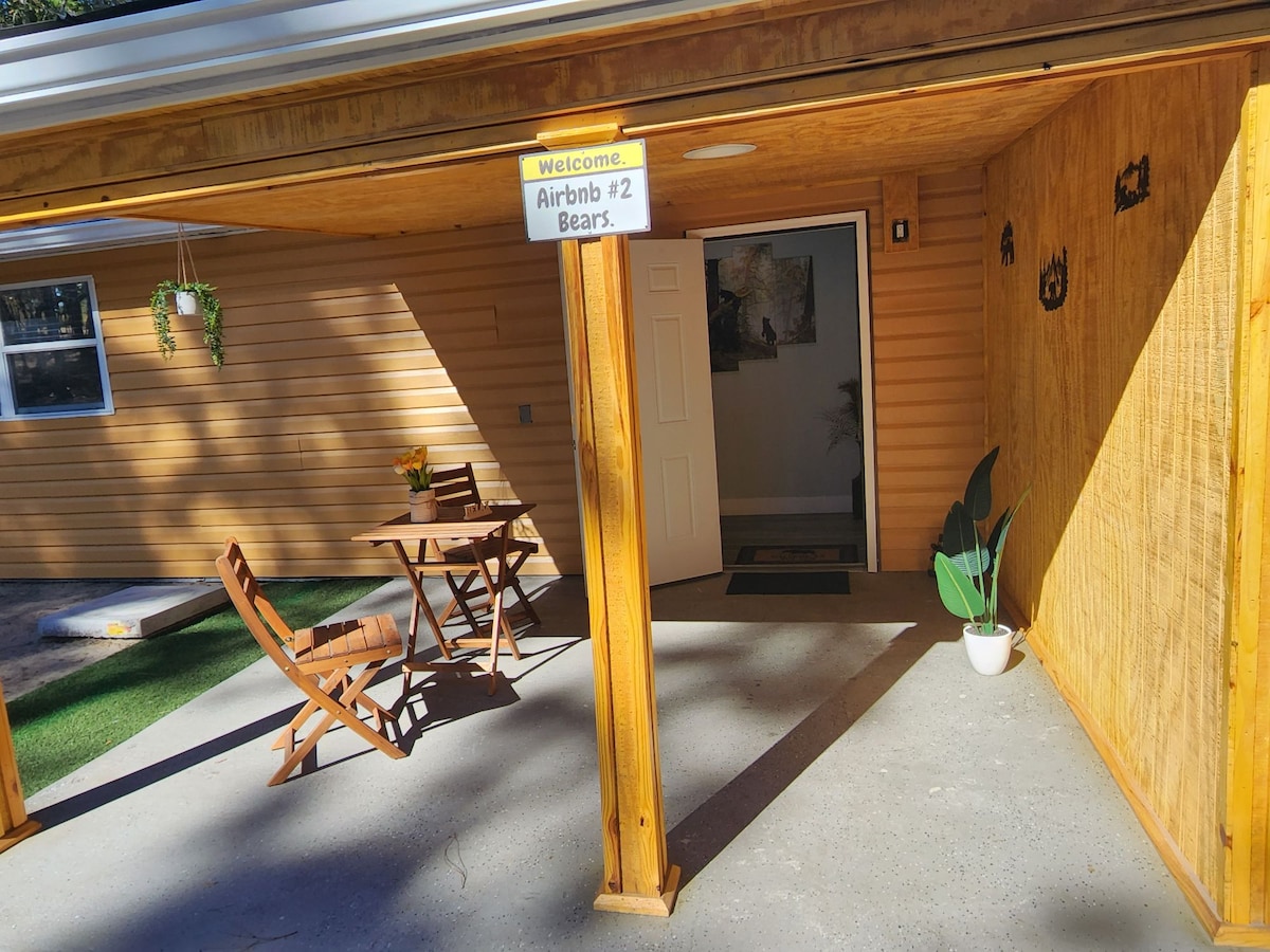 An entrance to a cabin features a covered porch with two wooden chairs and a small table, inviting relaxation. A welcome sign hangs above, while potted greenery adds a touch of nature. A door leads into the interior, framed by warm-colored walls.