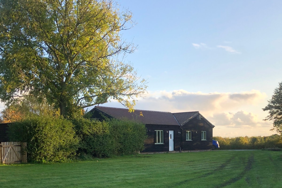 The single-storey barn is surrounded by a well-maintained green lawn and bordered by bushes and a tree. The rustic exterior features dark wood paneling, a shingled roof, and several windows that provide natural light. A pathway leads to the front door.