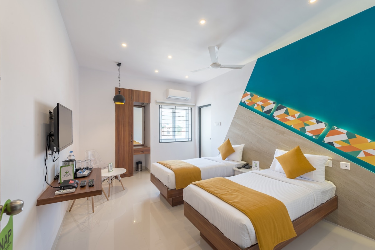 A modern studio room features two spacious beds with yellow accent pillows. A contemporary desk area is positioned near a window, allowing natural light. A wall adorned with geometric art in shades of teal and orange adds a vibrant touch, while a flat-screen TV is mounted for convenience.