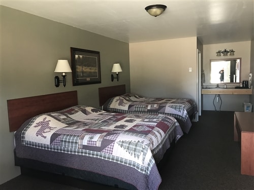 A refreshed room is presented featuring two spacious queen beds adorned with patterned quilts. Wall sconces provide soft lighting above each bed. A mirror and a wooden vanity area are visible in the background, creating an open, functional space.
