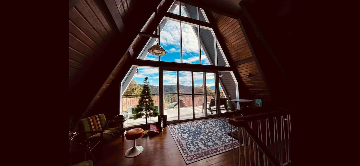A spacious living area features high wooden ceilings and large glass windows that offer expansive views of the mountains. A cozy rug lays on the wooden floor, and a Christmas tree adds a festive touch. Outdoor seating is visible through the glass doors, inviting relaxation.