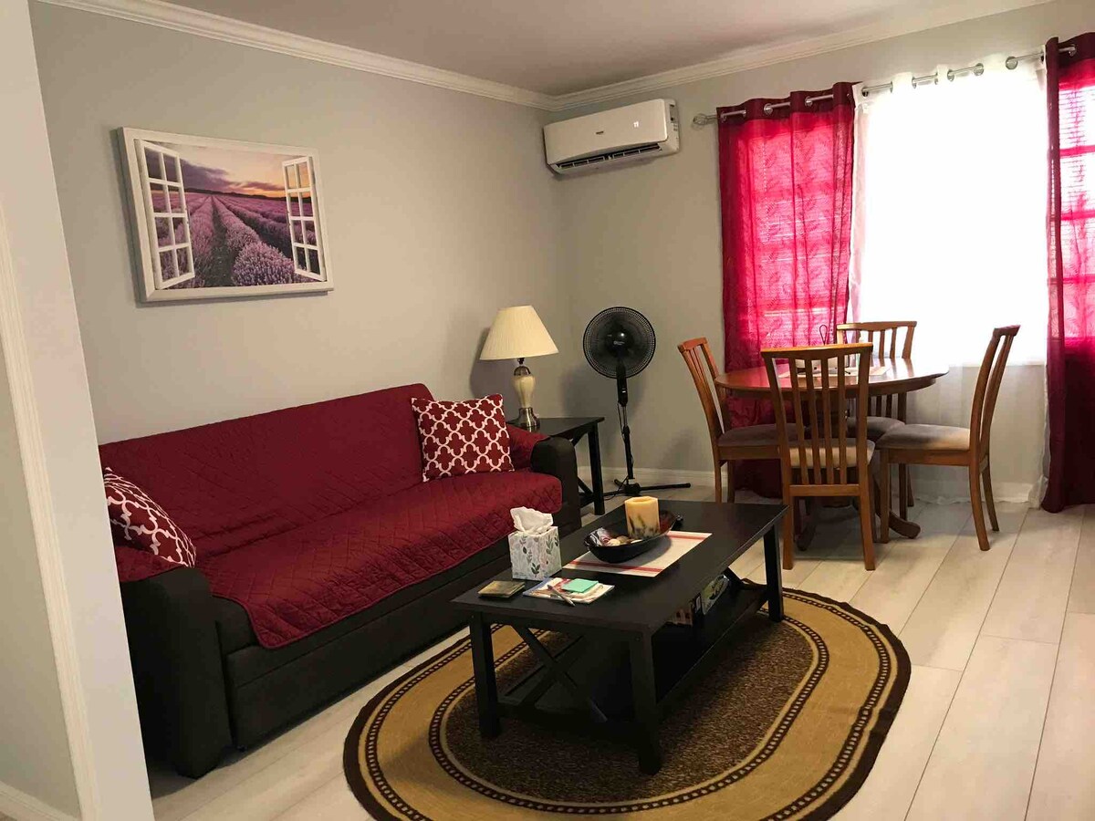 A comfortable living area features a dark sofa draped with a burgundy quilt and an adjacent lamp. A round dining table with four wooden chairs is positioned near a window, framed by red curtains. Air conditioning is present, and a fan enhances the space.