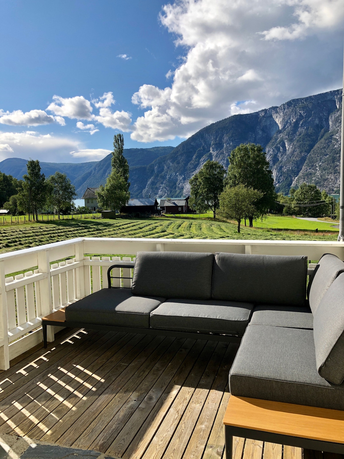 A comfortable outdoor seating area is set on a wooden deck, featuring a gray sectional sofa and a small side table. The backdrop includes rolling green hills and mountains, with a scenic view of nearby fields and trees reflecting the natural landscape.