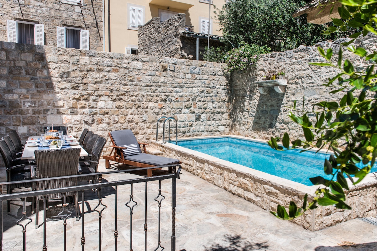 An intimate outdoor terrace features a heated pool surrounded by natural stone walls. A dining table with several chairs is set nearby, complemented by a sun lounger positioned beside the pool. Lush greenery adds a touch of nature to the inviting space.