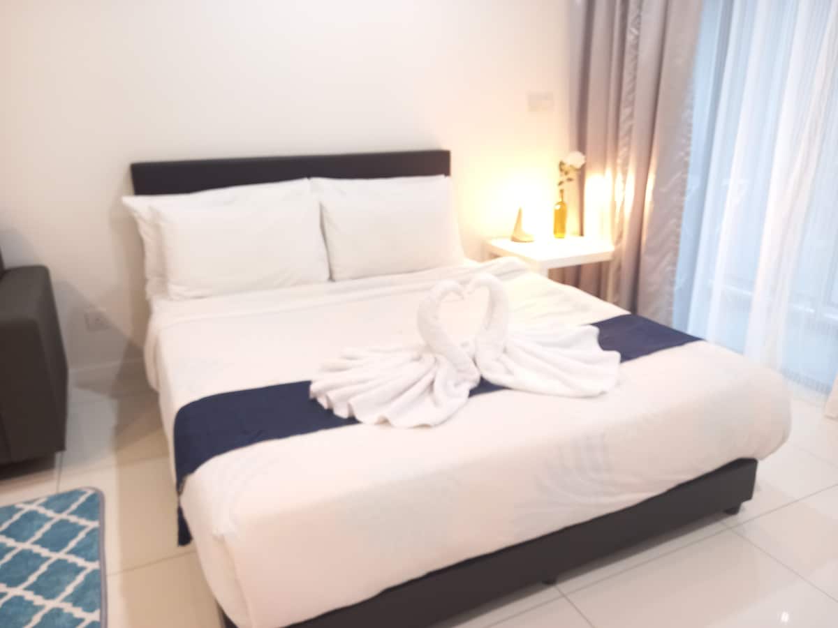 A cozy studio unit is displayed, featuring a spacious bed adorned with a towel arrangement resembling swans. Soft lighting from a bedside lamp highlights the clean, minimalist decor. Nearby, a window with light, flowing curtains allows natural light to brighten the space.
