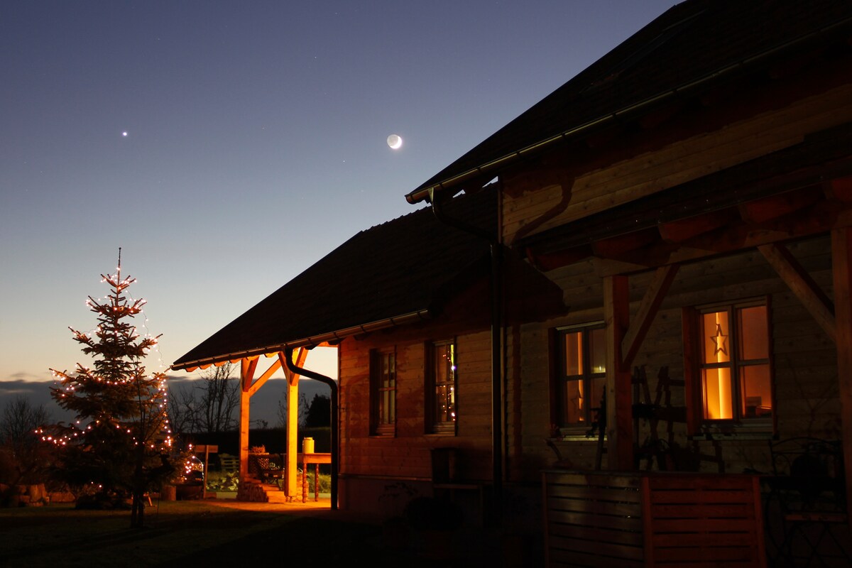A wooden house is softly illuminated at night, showcasing warm light emanating from the windows. A decorated Christmas tree stands nearby, adding festive charm. The sky features a crescent moon and stars above, complementing the peaceful ambiance of the surrounding landscape.