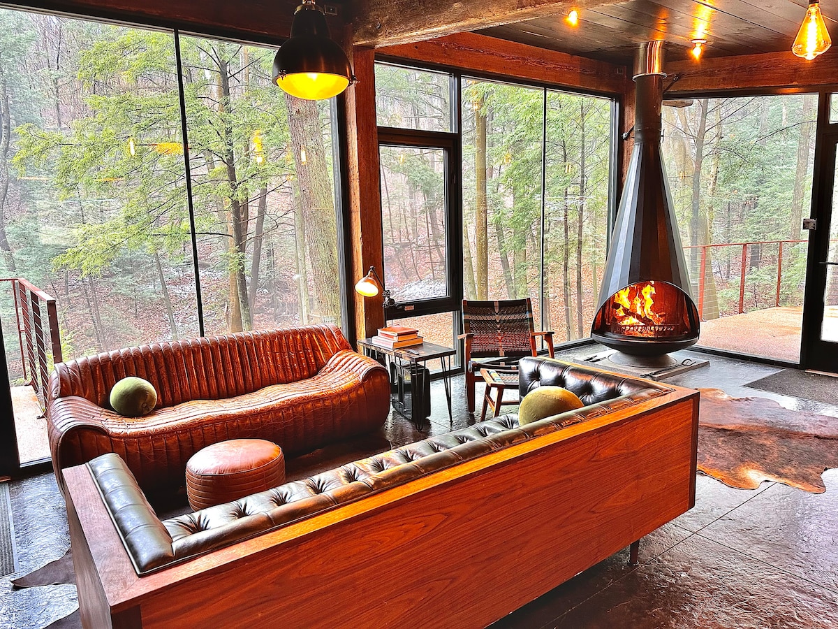 A spacious living area is highlighted by large floor-to-ceiling windows that provide views of the surrounding forest. A sleek, low-profile sectional sofa and a round ottoman are situated near a modern Malm fireplace, which adds warmth to the space. Two accent chairs complete the inviting setup.