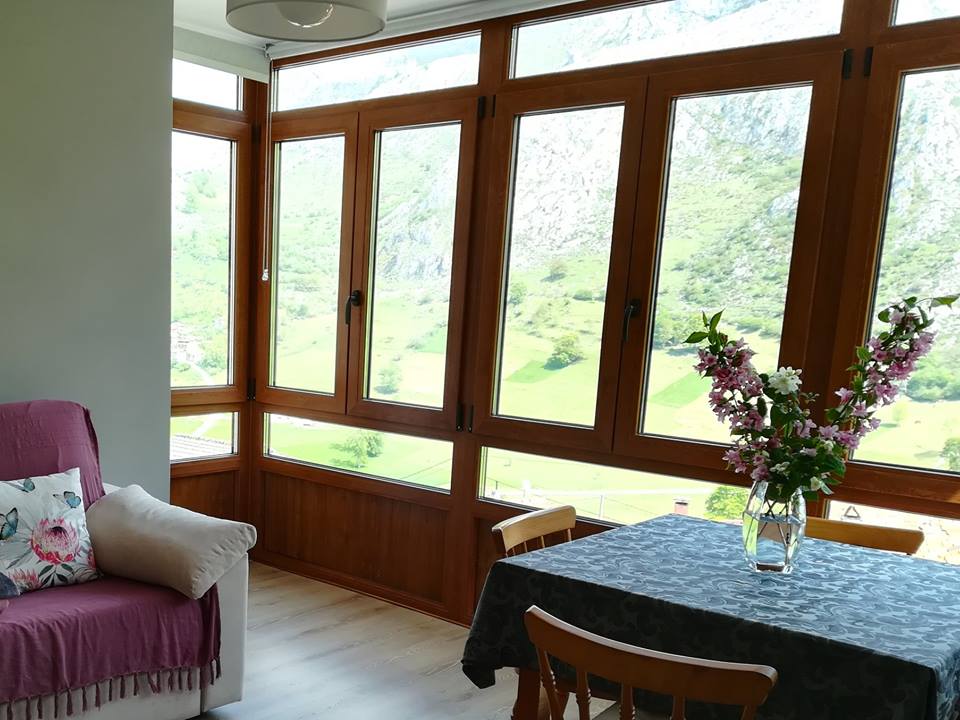 Large windows fill the space with natural light, offering views of lush green mountains. A wooden table is set with flowers in a vase, while a cozy seating area features a soft couch adorned with decorative pillows.